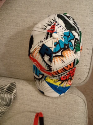 Färgglad keps med pop art-mönster - Säljer en riktigt cool keps med pop art-inspirerat tryck i flera färger som blått, grönt, orange, rött och gult. Kepsen har en böjd skärm och är täckt av grafiska texter och mönster som ger en unik och ungdomlig vibe. Perfekt för dig som vill sticka ut med din stil.