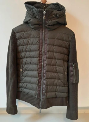 Mörkgrön pufferjacka Moncler med huva - Snygg mörkgrön pufferjacka från Moncler med stickade ärmar och quiltad kropp. Jackan har huva med snörning, tvåvägsdragkedja och ficka med dragkedja på ärmen. Materialmix av polyester och stickad ull ger en cool look. Moncler-logga på ärmen. Lappen inne i jackan är sönder som ja visar på en av bilderna men är annars i väldigt bra skick inga tecken på användning.