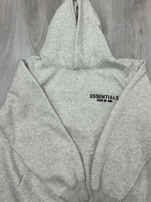 Essentials Hoodie - Den här hoodien är ljusgrå och har en stilren, minimalistisk design. På ryggen sitter den svarta texten ESSENTIALS FEAR OF GOD tryckt i ett tydligt och modernt typsnitt. Materialet är mjukt och tjockt, vilket ger ett bekvämt och värmande intryck. Den passar bra till vardagen.