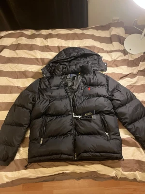 Svart pufferjacka från Polo Ralph Lauren - Svart pufferjacka från Polo Ralph Lauren med huva, flera dragkedjefickor och klassisk röd logga på bröstet. Jackan har en quiltad design och är tillverkad i polyester, perfekt för kalla dagar. Snygg och stilren look med normal passform.