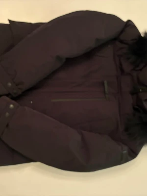 Svart parkajacka från Cecion Sweden - Parkajacka i svart från Cecion Sweden med huva och svart fuskpäls. Jackan har långa ärmar med ribbade muddar och tryckknappar, samt praktiska fickor framtill. Materialet är slitstark polyester och jackan har dragkedja framtill.