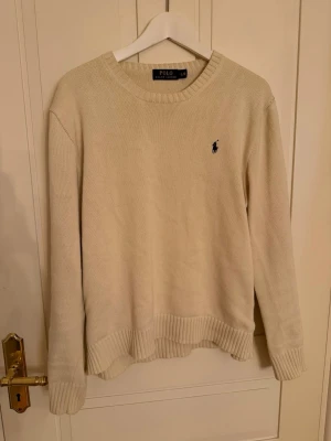 Beige stickad tröja från Polo Ralph Lauren - Snygg beige stickad tröja från Polo Ralph Lauren med klassisk rund hals och ribbade muddar. Tröjan har lång ärm och den ikoniska broderade loggan på bröstet. Perfekt för lager-på-lager under hösten och vintern. Storleken är L men passar M