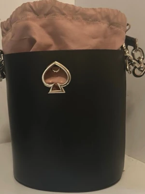 Svart bucket bag från Kate Spade - Säljer en svart bucket bag från Kate Spade med silvriga detaljer och logotyp framtill. Väskan har en rund form, axelrem och en beige insida med snörning upptill. Tillverkad i skinn och har en cool metallspade på framsidan. Perfekt för dig som gillar stilrena accessoarer.