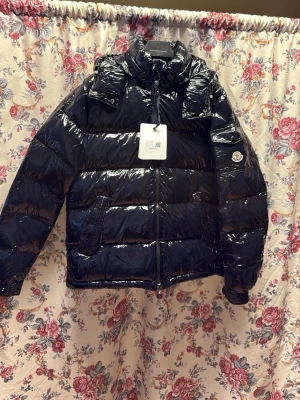 Maya moncler jacka - Den är svart ser mörkblå ut pga lampan säljer en glansig svart pufferjacka från Moncler med ikonisk logga på ärmen och coolt serietidningsfoder på insidan. Jackan har hög krage, dragkedja framtill och två sidofickor. Perfekt för dig som vill ha en snygg och varm jacka med street vibe.