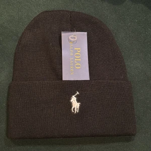 Svart stickad mössa från Polo Ralph Lauren - Snygg svart mössa från Polo Ralph Lauren med klassisk broderad logga framtill. Mössan är stickad och har en uppvikt kant för extra stilpoäng. Perfekt accessoar för att hålla värmen och se cool ut under kyliga dagar. Fråga vid minsta fundering.