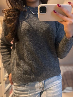 Mörkgrå stickad tröja - Mysig mörkgrå stickad tröja med rund hals och långa ärmar. Tröjan har en enkel och klassisk design med ribbade muddar vid ärmslut och nederkant. Perfekt att matcha med jeans för en chill look.