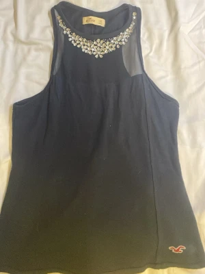 Svart blus med strass från Hollister - Supersnygg svart blus från Hollister med transparent mesh upptill och glittrande strassdetaljer runt halsen. Blusen är ärmlös och har en liten Hollister-logga broderad nertill. Perfekt för dig som vill ha något extra till outfiten.