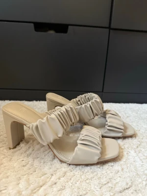 Beige sandaler med klack från H&M - Säljer ett par trendiga beige sandaler från H&M i storlek 36. Skorna har breda, rynkade remmar och fyrkantig tå. Klacken är stabil och med låg höjd, perfekt för dig som gillar stilrena detaljer. Materialet är syntet och de har en plattformsdesign.