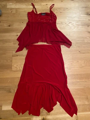 Röd paljettprydd linnetopp och kjol från Glitzy - Säljer en röd linnetopp från Glitzy med smala axelband och glittriga paljetter upptill. Toppen har ett flowigt, asymmetriskt fall. Matchande kjol följer med på köpet. Perfekt för dig som vill sticka ut med färg! Sommar staple!!
