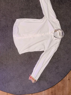 Vit Burberry skjorta med rutiga detaljer - Säljer en stilren vit skjorta från Burberry Brit i storlek S. Skjortan har klassiska rutiga detaljer på insidan av kragen och vid ärmsluten, samt knappar framtill. Perfekt för dig som gillar ikoniska designer och vill ha en clean look.
