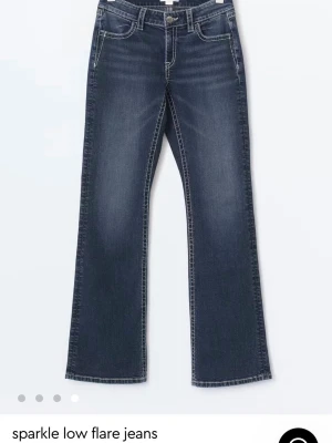 Blå flare jeans med broderade fickor - Snygga blå jeans med låg midja och flare-ben. Baktill finns broderade fickor med glittriga detaljer som ger extra attityd. Jeansen har klassisk femficksdesign och kontrastsömmar. Perfekta för dig som vill sticka ut lite extra.