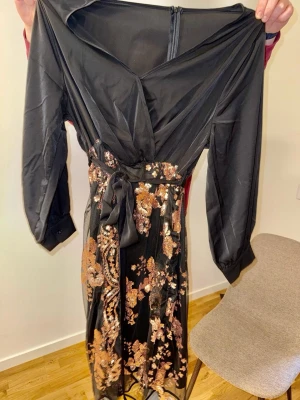 Svart långklänning med blommiga paljetter - Svart långklänning med v-ringad omlott upptill och långa ärmar. Kjolen har blommigt mönster i guld och kopparfärgade paljetter, samt ett knytband i midjan. Klänningen har dragkedja i ryggen och är tillverkad i ett tunt, glansigt material. Glitterklänning storlek 46/48 samt passar 48/50.