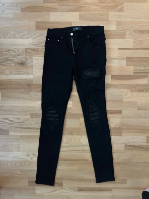 Svarta skinny jeans från Amiri - Svarta jeans från Amiri i skinny fit med slitna detaljer och hål på båda benen. Klassisk femficksmodell med silverfärgad knapp och dragkedja framtill. Materialet är jeans och byxorna har en modern, tajt passform som sitter snyggt.