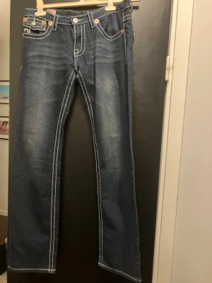 True Religion Joey Super T jeans blå - Säljer ett par blå True Religion Joey Super T jeans med kontrasterande vit och röd söm, klassiska fickor med lock och emblem på baksidan. Bootcut-modell med grova stickningar och detaljerade knappar. Märkespatch med logga och gitarrmotiv bak.