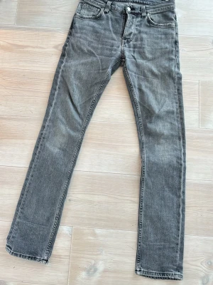 Grå jeans från Nudie Jeans, 28/32 - Säljer ett par grå jeans från Nudie Jeans i storlek 28/32. De har klassisk femficksdesign, svarta broderade detaljer på bakfickorna och svart patch bak i midjan. Jeansen är raka i modellen och tillverkade i slitstarkt bomullsmaterial. Sitter som w27