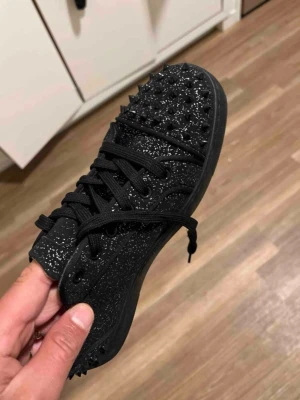 Svarta sneakers med nitar och glitter - Säljer ett par svarta sneakers med coola svarta nitar på tån och glittereffekt över hela skon. Skorna har snörning och en unik röd sula med grafiskt mönster. Perfekt för dig som vill sticka ut med din stil.