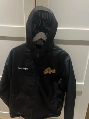 Palm Angels svart vindjacka med nalle - Säljer en svart vindjacka från Palm Angels med huva och dragkedja. Jackan har ett broderat nallemärke på bröstet och Palm Angels-logga i vitt. Insidan är fodrad med stort Palm Angels-tryck. Perfekt för dig som vill ha streetwear-vibe.