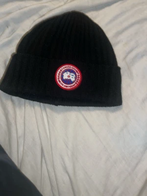 Svart ribbad mössa från Canada Goose - Säljer en svart ribbad mössa från Canada Goose med klassisk logga framtill. Mössan är tillverkad i 65% bomull och 35% polyester och har en bred uppvikt kant för extra stilpoäng.