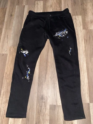 Svarta mjukisbyxor med tryck och färgstänk - Svarta mjukisbyxor från Excite med coolt tryck på ena låret och färgstänk i blått, gult och vitt. Byxorna har snörning i midjan och en slim passform. Perfekta för chill dagar och streetwear-stil.