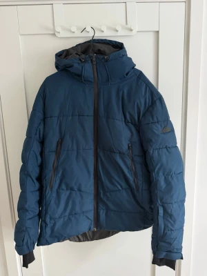 Blå McKinley pufferjacka XL - Blå pufferjacka från McKinley i storlek XL. Jackan har huva, två fickor med dragkedja och hel dragkedja framtill. Materialet är syntet och jackan har en quiltad look med långärmade ärmar. Perfekt för kalla dagar och outdoor.