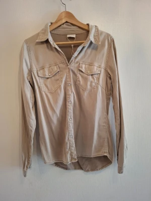 Beige skjorta med knappar från Vila - Snygg beige skjorta från Vila i bomull med klassisk krage och två bröstfickor med lock. Skjortan har långa ärmar och tryckknappar framtill. Passformen är normal och materialet känns mjukt och bekvämt. Perfekt för dig som gillar stilrena och enkla plagg.