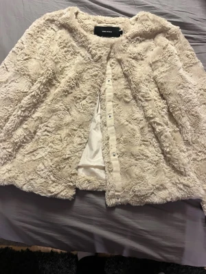 Beige pälsjacka från Vero Moda - Mysig beige pälsjacka från Vero Moda i storlek M. Jackan har ett fluffigt och mjukt material, rund hals och knappar framtill. Perfekt för dig som vill ha en varm och stylish look under kyliga dagar.
