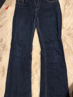 Bootcut jeans mörkblå W27 L32 - Mörkblå bootcut jeans med klassisk orange söm och fem fickor. Jeansen har en snygg bootcut-passform som ger lite extra vidd vid benslutet. Materialet är jeans och färgen är djup mörkblå, perfekt för en chill och stilren look.dem är uppsydda och jag är 156 cm