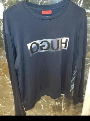 Mörkblå långärmad tröja från Hugo - Snygg mörkblå långärmad tröja från Hugo med stor logotyp i silver och svart på bröstet. Tröjan har rund hals och är gjord i mjuk bomull, perfekt för en chill och stilren look. Passar dig som gillar klassiska streetwear-vibbar.