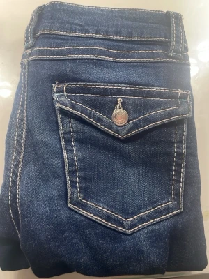 Blå bootcut jeans med ficklock - Snygga mörkblå jeans med bootcut-snitt och markerade vita sömmar. Baksidan har ett ficklock med knappdetalj som ger extra stil. Säljer pga för små.