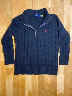 Svart stickad tröja Polo Ralph Lauren - Svart kabelstickad tröja från Polo Ralph Lauren med halv dragkedja och röd broderad logga på bröstet. Tröjan har hög krage, lång ärm och är gjord i ett mjukt stickat material. Perfekt för kyliga dagar och snygg till jeans.