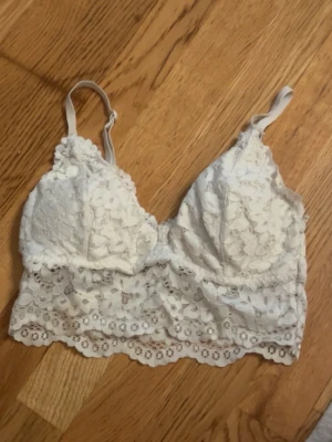 Vit spets topp - Supersöt vit bralette topp med blommig spets och justerbara axelband. Toppen har en bred spetskant nedtill och har ilägg på insidan för extra komfort. 