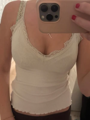 Ribbad vit linnetopp med spets - Supersöt ribbad vit linnetopp med spetsdetaljer längs v-ringningen och axelbanden. Toppen är figurnära och har en lätt vågig kant nertill. Perfekt att styla med jeans eller kjol för en fräsch look.