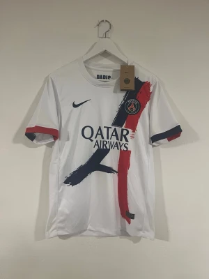 PSG Nike vit fotbollströja S - Säljer en officiell Paris Saint-Germain fotbollströja från Nike i storlek S. Tröjan är vit med marinblå och röda penseldrag, PSG-logga på bröstet och Qatar Airways-tryck. Ärmarna har blå och röda detaljer. Materialet är lätt polyester, perfekt för fotboll.