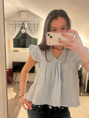 Ljusblå blus med volangärmar - Supersöt ljusblå blus med v-ringning och volangdetaljer på ärmarna. Blusen har en lös passform och är tillverkad i ett tunt, mjukt bomullstyg. Perfekt för vår och sommar när du vill ha något luftigt och stilrent.