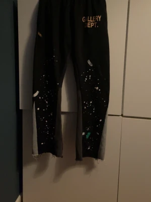 Svarta mjukisbyxor från Gallery Dept. - Svarta mjukisbyxor från Gallery Dept. med färgstänk och tryckt logga på framsidan. Byxorna har grå paneler längs benen och råa, fransiga benslut. Tillverkade i 100% bomull för skön känsla. Perfekt för dig som gillar streetwear och unik design.