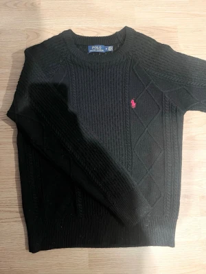 Svart stickad tröja Polo Ralph Lauren - Svart stickad tröja från Polo Ralph Lauren i storlek M. Tröjan har ett snyggt kabelstickat mönster med diskreta rutor och en röd broderad logga på bröstet. Rund hals och ribbade muddar vid ärmslut och nederkant. Perfekt för dig som gillar klassisk stil.