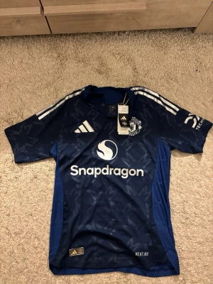 Manchester united fotbollsjersey helt ny  - Säljer en blå Manchester United tröja från adidas i storlek S. Tröjan har ett mönster med ljusblå detaljer, vita ränder på axlarna och klubbens logga på bröstet. Materialet är lätt och andas, perfekt för fotboll eller träning. Slim fit-modell med snygg design. Helt ny! Passar folk mellan 170-185. Tveka inte att höra av er vid frågor eller funderingar🤗