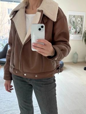 Brun teddyjacka från Amisu - Säljer en brun teddyjacka från Amisu med mjukt, vitt foder och stor krage. Jackan har dragkedja, sidofickor och detaljer med knappar och bälte vid midjan. Materialet är polyester och jackan har en loose passform som ger en chill vibe.