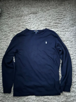 Marinblå långärmad tröja Polo Ralph Lauren - Snygg marinblå långärmad tröja från Polo Ralph Lauren i storlek XL i barnstorlek, som liknar S i herrstorlek. Tröjan har rund hals och klassisk broderad logga i vitt på bröstet. Tillverkad i mjuk bomull som är skön mot huden och passar perfekt till jeans eller chinos.
