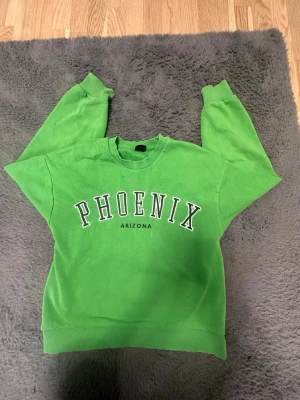 Grön sweatshirt med PHOENIX-tryck - Cool grön sweatshirt med PHOENIX ARIZONA-tryck på bröstet. Tröjan har rund hals, ribbade muddar vid ärmslut och nederkant samt långa ärmar. Perfekt för dig som gillar streetstyle och vill sticka ut med färg. Materialet känns som mjuk bomull.
