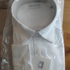 Vit skjorta från These Glory Days - Klassisk vit skjorta från These Glory Days med snygg krage och knäppning framtill. Skjortan är tillverkad i bomull och har långa ärmar med knappar vid manschetten. Perfekt för en clean och stilren look.  Bröst 120cm Midja  115cm Hals 43/44 cm Arm 68 cm