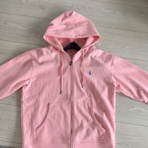 Rosa hoodie från Polo Ralph Lauren - Säljer en ljusrosa hoodie från Polo Ralph Lauren med dragkedja och huva. Tröjan har långa ärmar, snörning i huvan och en blå broderad logga på bröstet. Tillverkad i mjuk bomull och har praktiska fickor framtill.