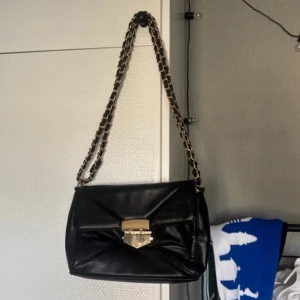 Svart axelväska med guldkedja - Snygg svart axelväska i skinnimitation med guldfärgad kedjerem och dekorativt lås framtill. Väskan har quiltad framsida, dragkedjeficka på baksidan och rymligt innerfack. Perfekt för dig som vill ha en stilren och trendig väska med lyxig känsla.