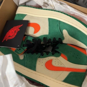 Nike Air Jordan 1 gröna och orange - Nike Air Jordan 1 sneakers i en riktigt fräsch färgkombo med grönt mocka, beige paneler och orange swoosh. Klassisk siluett med svart snörning och vit sula. Perfekta för dig som vill sticka ut med dina sneakers.