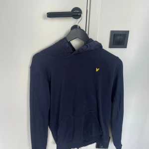 Mörkblå hoodie från Lyle & Scott - Säljer en mörkblå hoodie från Lyle & Scott med klassisk gul logga på bröstet. Tröjan har huva med snörning och en stor magficka framtill. Perfekt för dig som gillar en enkel och stilren look. Storlek XS-S