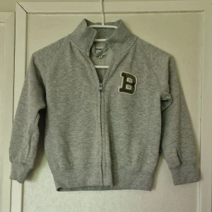 Grå ziptröja med camo-detaljer - Grå ziptröja från Barracuda med hög krage och ett B-märke på bröstet. Ärmarna har camo-lappar på armbågarna för extra stilpoäng. Ribbade muddar och hel dragkedja framtill. Tröjan är nopprig, haken håller på att lossna på ena sidan och det finns två mindre fläckar som inte syns om man inte granskar den (se bilder). Jag kan inte tyda vad det är för material.