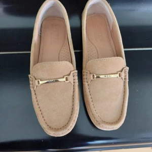 Beige mockaloafers från Ralph Lauren - Snygga loafers i beige mocka från Ralph Lauren med guldfärgad metall-detalj framtill där det står RLL. Skorna har klassisk rund tå och platt sula med gummiknoppar baktill för extra grepp. Perfekta för en stilren och avslappnad look. Storlek 36 men det är en väldigt smalmodell. 