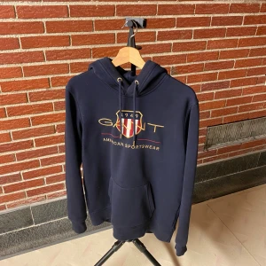 Mörkblå hoodie från Gant med brodyr - Säljer en mörkblå hoodie från Gant med broderad logga och sköld på bröstet. Tröjan har huva med snörning, känguruficka och långa ärmar. Perfekt för dig som gillar klassisk och sportig stil.