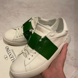 Valentino Garavani Open sneakers vit/grön - Snygga Valentino Garavani Open sneakers i vitt skinn med en bred grön läderdetalj över snörningen. Klassisk rund tå, platt sula och diskret logga i guld på plösen. Kommer med originalkartong och dustbag. Perfekta för dig som gillar exklusiva och trendiga sneakers. Pris kan sänkas vid snabbaffär