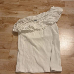 Vit offshoulder topp med volang Kappahl - Superfin vit offshoulder topp från Kappahl med bred volangkant i broderad spets. Toppen är ribbad och har en ärmlös, asymmetrisk design som ger en trendig look. Perfekt för dig som gillar romantiska detaljer och vill sticka ut lite extra.